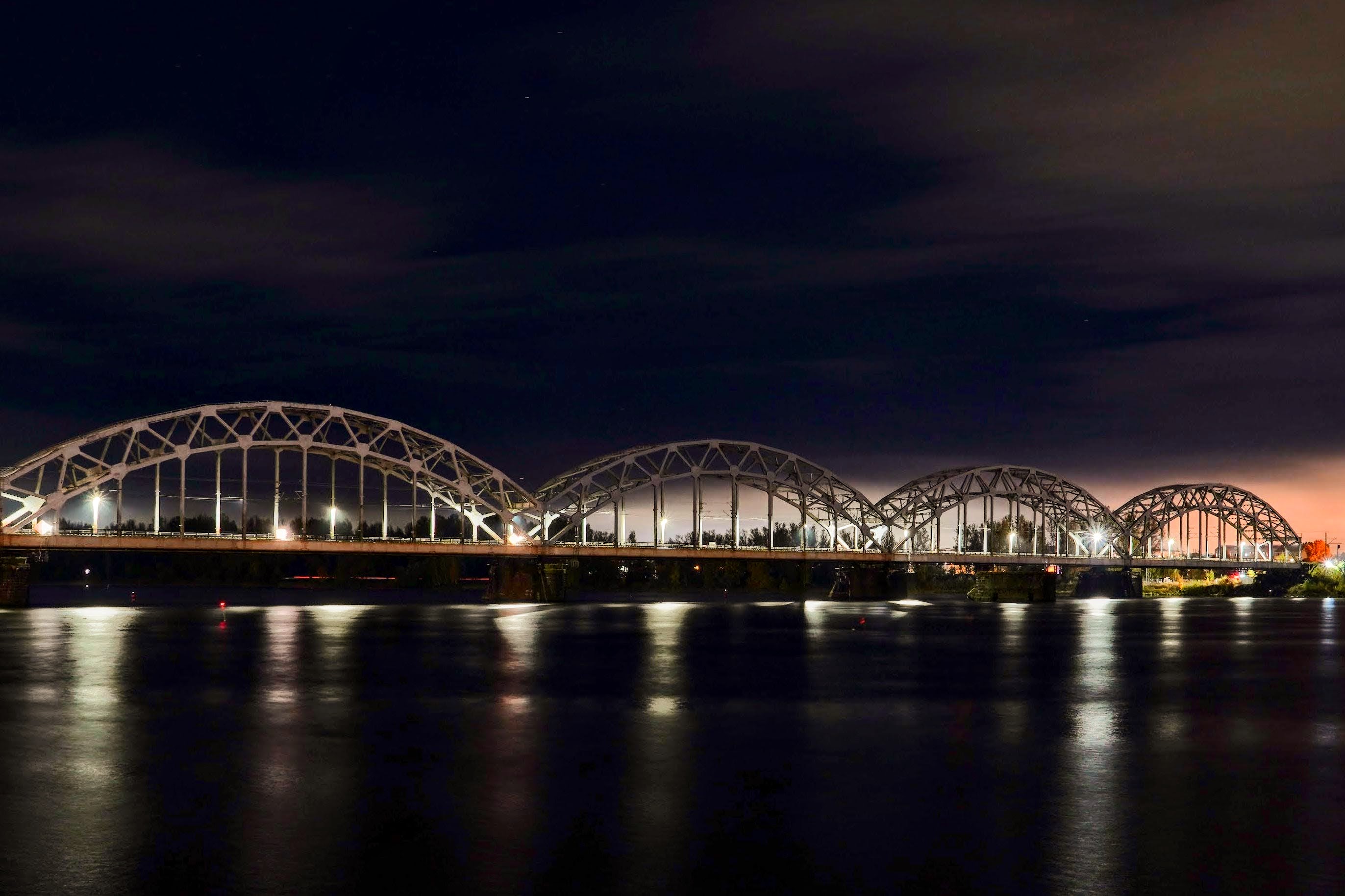 Die eiserne Brücke spannt eine helle Verbindung über den dunklen Fluss in Riga