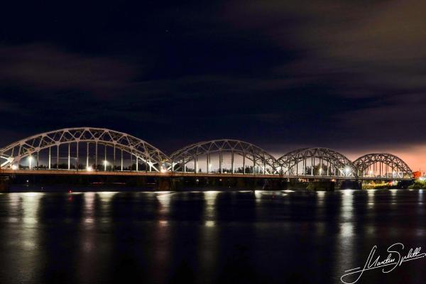 Die eiserne Brücke spannt eine helle Verbindung über den dunklen Fluss in Riga